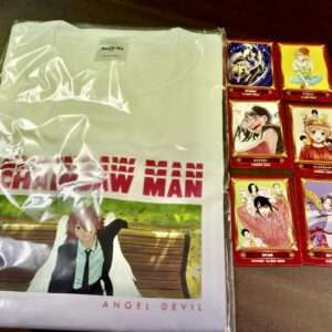 White packaged T‑shirt featuring Angel Devil artwork beside six red-framed anime cards on the right, on a dark table.