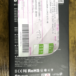 Black rectangular product box with a pink shipping label affixed diagonally across the front, barcode and small text visible beneath the label, set on a wooden surface