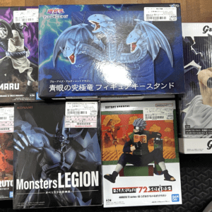 Assorted anime action figure boxes on a wooden floor, including Orochimaru, a Blue-Eyes Ultimate Dragon figure, Grandista Sasuke, Uzumaki Naruto, Monsters LEGION figure, and Naruto 72 Series boxes