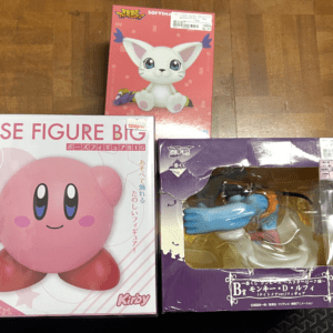 Three boxed character figures on a wooden floor: a large pink Kirby figure box, a small pink box with a white cat-like character, and a purple box with a figure visible through a window.
