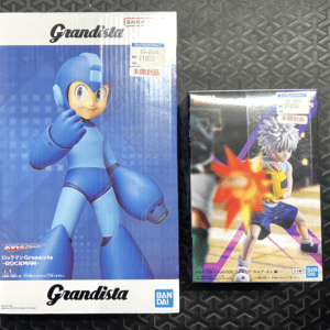 Two boxed action figures on a dark textured surface: a large blue Mega Man Grandista box on the left and a Hunter x Hunter figure box on the right with price stickers visible on both boxes.