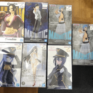 Boxed anime figures arranged on a wooden surface: multiple Bandai Grandista and MAXMATIC figures in their boxes, featuring stylized characters with bold costumes and poses in a 2x4 grid layout.