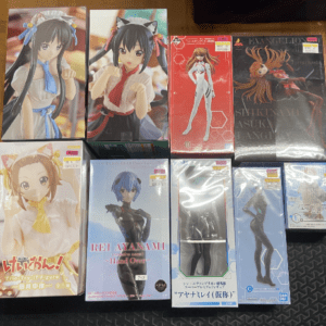 Collection of boxed anime figure boxes laid out on a wooden floor, showing colorful character outfits and packaging from multiple series.