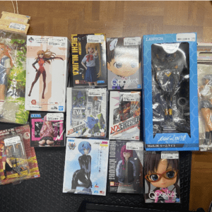 Assorted anime action figures and boxed collectibles scattered on a wooden floor, including Nendoroid- and Qposket-style boxes in various sizes and colors.  Articulos Japan (Post Box Japan)