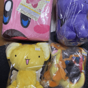 Four plush stuffed toys in sealed plastic bags on a black surface: pink round character, purple character with a cap, yellow bear-like figure, and an orange dragon-like toy with blue accents.