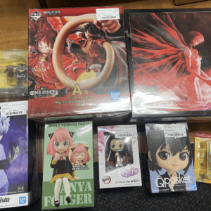 A collection of anime figure boxes and small figures laid out on a wooden floor, including One Piece, Evangelion, Grandista, and Qposket toys.