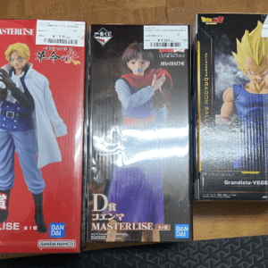 Three anime figure boxes on a wooden floor: left red B賞 Masterline, middle dark boxer-like figure, right Grandista-VEGETA with yellow hair or Super Saiyan Vegito design.