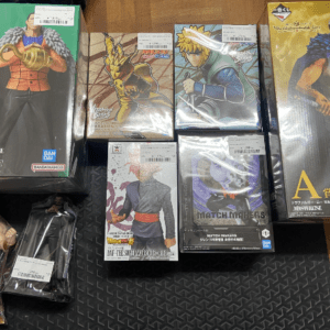 Assorted boxed anime action figures arranged on a wooden floor, including a large green-packaged figure on the left and a tall blue-waisted figure on the right, with several smaller boxes in the center. Brand logos visible (Bandai).
