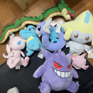 Pile of colorful plush Pokémon toys on a wooden floor, including a purple Gengar and several pastel characters.",