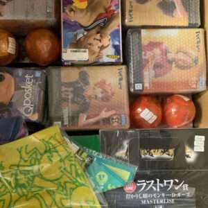 Box filled with stacked anime figurine boxes; colorful covers feature characters, with two orange balls and a yellow-green cloth in the corner.