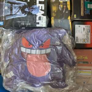Purple Gengar plush toy in a plastic bag inside a cardboard box among boxed anime figures and collectibles.