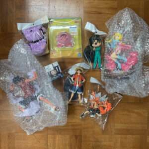 Assorted anime figures and accessories, still in plastic packaging and bubble wrap, arranged on a wooden floor.