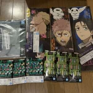 Spread of anime posters and merchandise on a wooden floor, featuring three tall character posters with bold faces and a row of character pins/keychains in green and black packaging nearby.