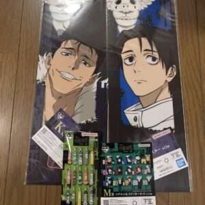 Pair of vertical anime posters featuring two male characters with a white winged skull motif above, laid on a wooden floor, with bundled keychains and magnets in front (Bandai Namco).