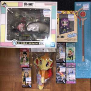 Display of hobby figurines in colorful boxed packaging on a wooden floor, with plush toy and various character figures labeled with price tags (Japanese store setting)