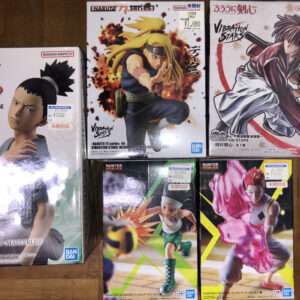 Six boxed anime figures arranged on a wooden floor: left is a tall white Naruto Masterlise box featuring a character portrait; top center is a Deidara/ Naruto Vibrations Stars figure in an action pose; top right a Vibrations Stars box showing a swordsman character; bottom left is a Hunter x Hunter figure of Gon Freecss in a dynamic stance; bottom right is a Hunter x Hunter figure of Hisoka with a pink aura.