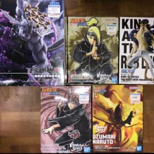 Row of boxed anime figures on a wooden floor: large left box with a gray-purple creature, middle box shows Naruto in a black robe with hand seal, right box features a tall character in white pants and a dark vest. Bottom row shows two Naruto figures in dynamic poses.