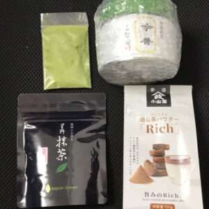 Assorted Japanese tea products: a small bag of yellow-green powder, a white-and-green tin, a black resealable pouch labeled Japan Green, and a white pouch labeled Rich with a cookies-and-cup image on the front on a dark surface.  Articulos Japan (Post Box Japan)