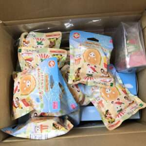 Open cardboard box filled with colorful snack or toy packages featuring cute animal characters in blue and cream wrappers, laid haphazardly inside a box.