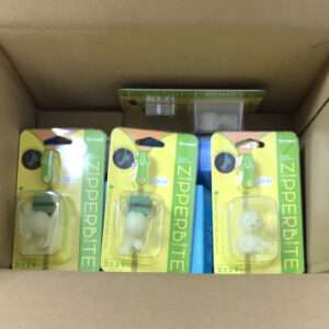 Three yellow-green Zipperbite product boxes in a brown cardboard carton, each showing a clear plastic window with a white, bear-shaped item inside.