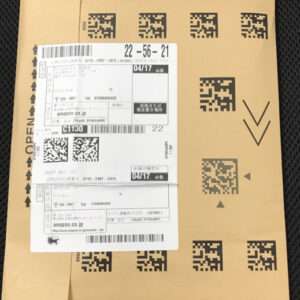 Brown cardboard shipping box with multiple overlapping white shipping labels featuring barcodes and QR codes, including Japanese text, lying on a dark textured surface.  Articulos Japan (Post Box Japan)