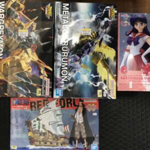 Collection of five boxed anime figures/model kits on a dark surface: two large Digimon boxes labeled WarGreymon and MetalGarurumon, a pink boxed figure featuring a female anime character, and a One Piece ship diorama box in the foreground.