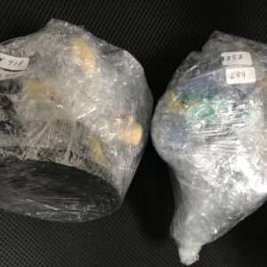 Two plastic-wrapped bundles sitting on a dark perforated surface, each with handwritten price stickers showing multiple numbers (left and right bundles).  Articulos Japan (Post Box Japan)