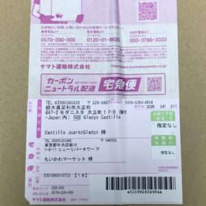 Pink Japanese delivery label on a cardboard box with a barcode, tracking numbers, and recipient address text (Japanese).