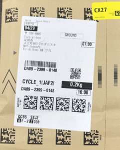 Shipping label on a cardboard box with 'GROUND', date 04/29, weight 0.2 kg, time 16:00, and multiple barcodes/QR codes visible.  Articulos Japan (Post Box Japan)