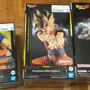 Three Dragon Ball Z action figure boxes on a wooden floor: Mecha Makers Goku on the left, Grandista Son Goku II in the center, and a Super Saiyan figure in blue on the right.