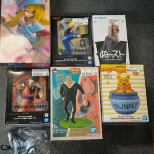 Row of anime figure boxes on a dark carpet: Zamasu-like character on left, затем Match Makers boxes in center, Noodle Stopper Pooh box on right, plus a Dragon Ball figure and Revive Moment box nearby.