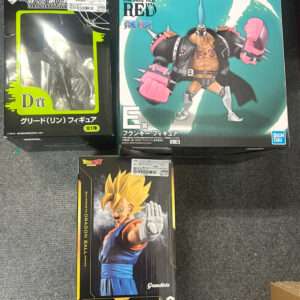 Three boxed anime figures on a dark carpet: a neon-green/black Greeed figure box on the left, a large One Piece Film Red figure box on the right, and a Dragon Ball Z Vegito figure box in front.