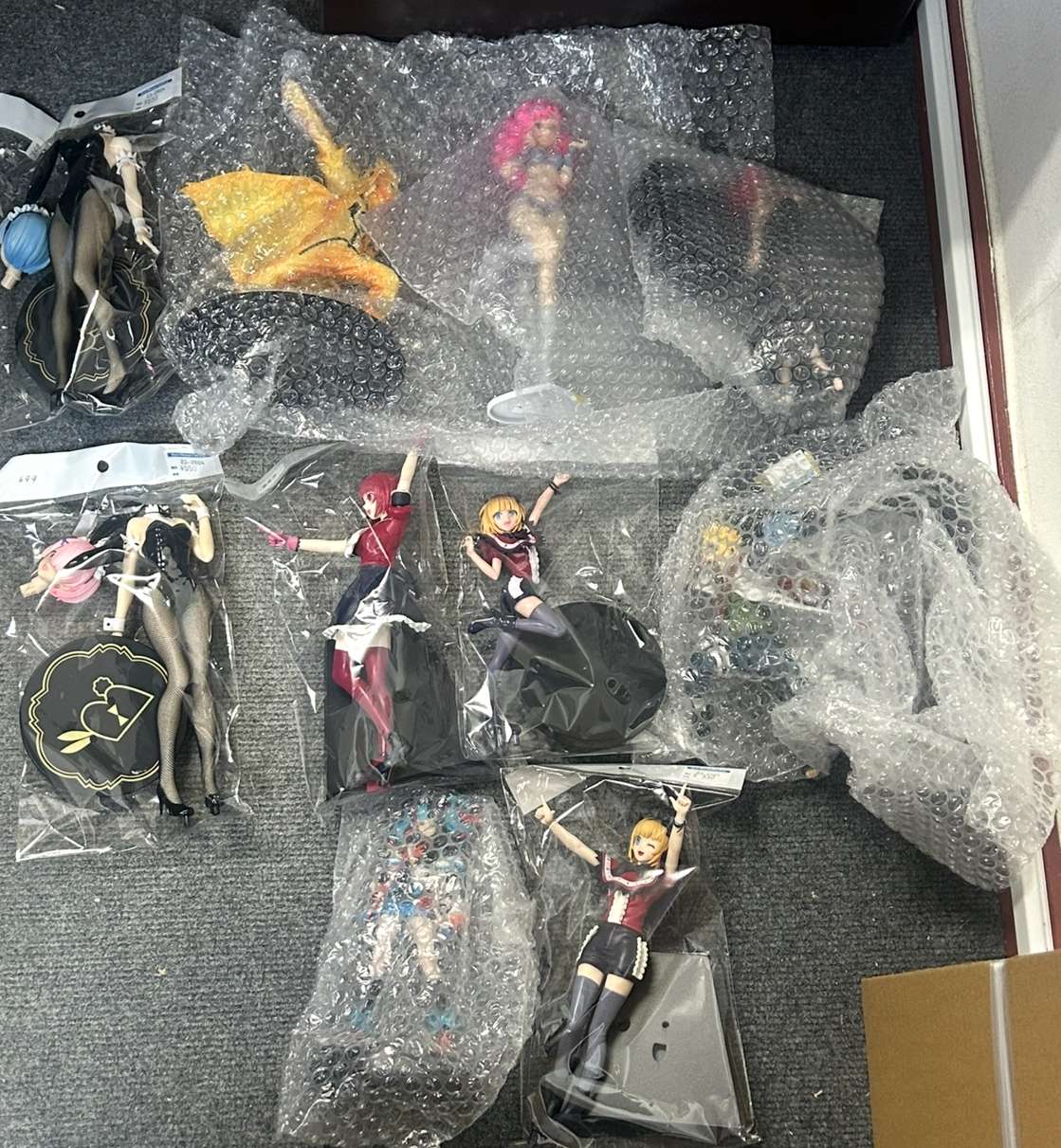 Img_9329 - Articulos Japan (post Box Japan) Collection of anime figurines in plastic bags and bubble wrap laid out on a gray carpeted floor, ready for packaging or display. Articulos Japan (Post Box Japan)