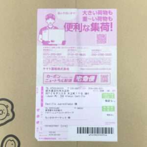 Pink Japanese delivery label on a cardboard box, showing a cartoon courier holding a box, QR code, and contact numbers.
