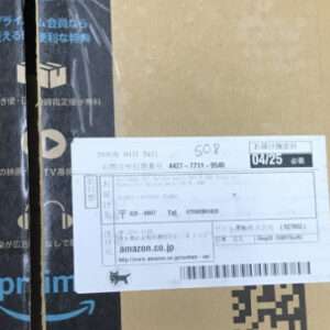 Cardboard Amazon Prime box with a white shipping label in the center, showing a barcode and Japanese text; blue Prime branding on the left and a small QR code on the right.
