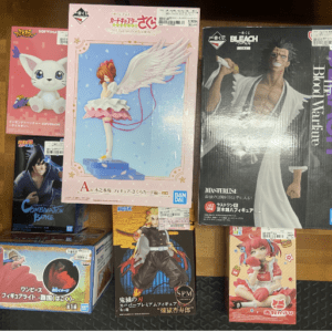 Assorted boxed anime figures arranged on a wooden shelf, including a pink winged girl center and a Bleach statue on the right.
