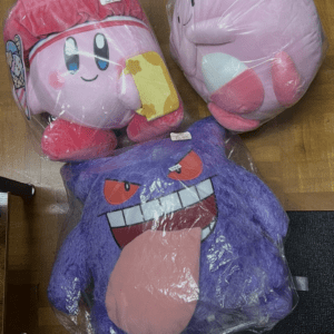 Three large plush toys in plastic wrap on a wooden floor: two pink Kirby‑like characters and a purple monster with a protruding tongue.