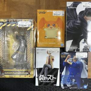Five boxed anime figures on a wooden floor: a large figure in protective packaging on the left and four other character figure boxes on the right.