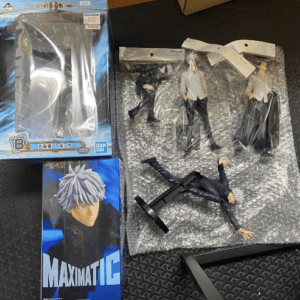 Assorted anime figures and boxes on a dark, textured surface: a boxed Maximatic figure on the left, bubble-wrapped figures in the center-right, and a blue Maximatic box featuring a character with white hair.