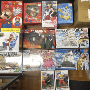 Assorted anime and video game character figure boxes arranged on a wooden floor, including Mario and One Piece boxes to the left and right edges