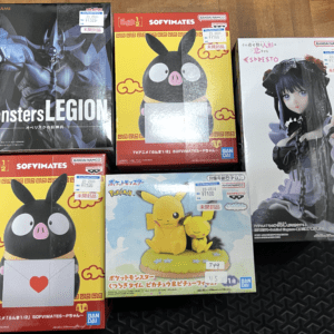 A collection of boxed anime figurines on a wooden floor: top left 'Monsters LEGION' demon figure, top middle a yellow Sofvimates pig box, top right a tall gothic girl figure, bottom left another Sofvimates pig box with a heart envelope, and bottom center a Pikachu figure displayed with Eevee on a grassy sky-blue background.