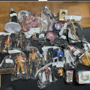 Assorted anime action figures in plastic bags spread across a dark mat on a wooden floor, many characters and packaging visible.