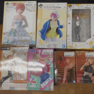 Row of boxed anime figures on the floor; leftmost shows a woman in a white wedding dress, center a pink-haired character in a yellow outfit, right a figure in a clear window box apartment style outfit.  Articulos Japan (Post Box Japan)