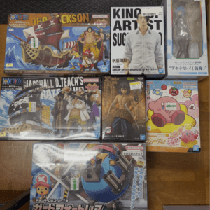 Collection of Bandai model kits and figures laid out on a table, including One Piece ship set and a King of Artist Suguru Geto figure box with other anime boxes nearby.