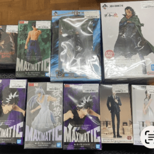 Collection of boxed anime figures laid out on a wooden floor, including Maximatic boxes and a large white-box figure with a cloaked character on the right.