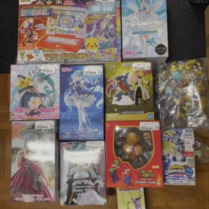 Top-down view of a collection of boxed anime figures and toys spread out on a wooden floor (Hatsune Miku, Mario, Pokemon items visible).