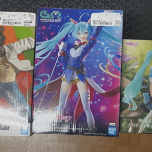 Three boxed anime figures on a wooden table: left is a Grandista figure with a character wearing a white jacket and a chainsaw for a head; center is Hatsune Miku in a blue stage outfit; right shows a turquoise-haired girl in a teal maid-style dress in a garden setting.