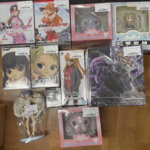 Assorted anime figure boxes arranged on a wooden floor, featuring characters in colorful packaging and large display boxes that hint at different series and franchises.