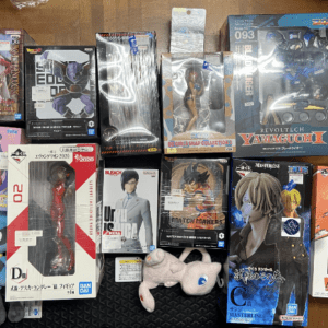 A collection of boxed anime figures arranged on a dark floor mat, featuring character figures in various packaging styles and sizes.