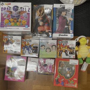 Assorted boxed anime action figures on a wooden floor, including Dragon Ball Z and Grandista figures, with a yellow plush character on the right.  Articulos Japan (Post Box Japan)